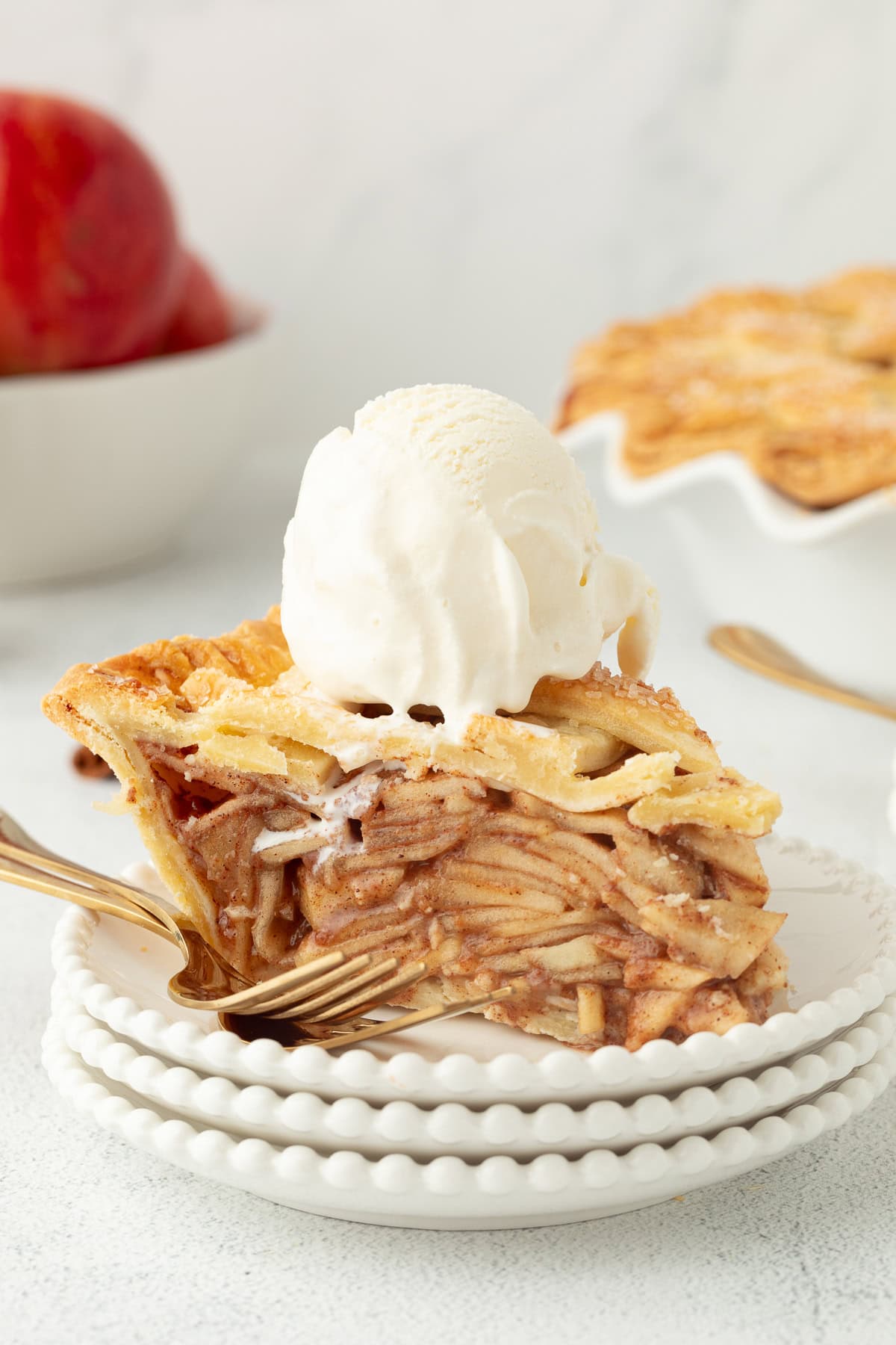 A slice of apple pie topped with a scoop of ice cream on a white plate with the rest of the pie in the background.