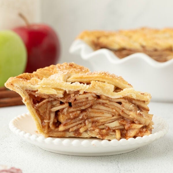 A slice of apple pie on a white plate with the rest of the pie in the background.