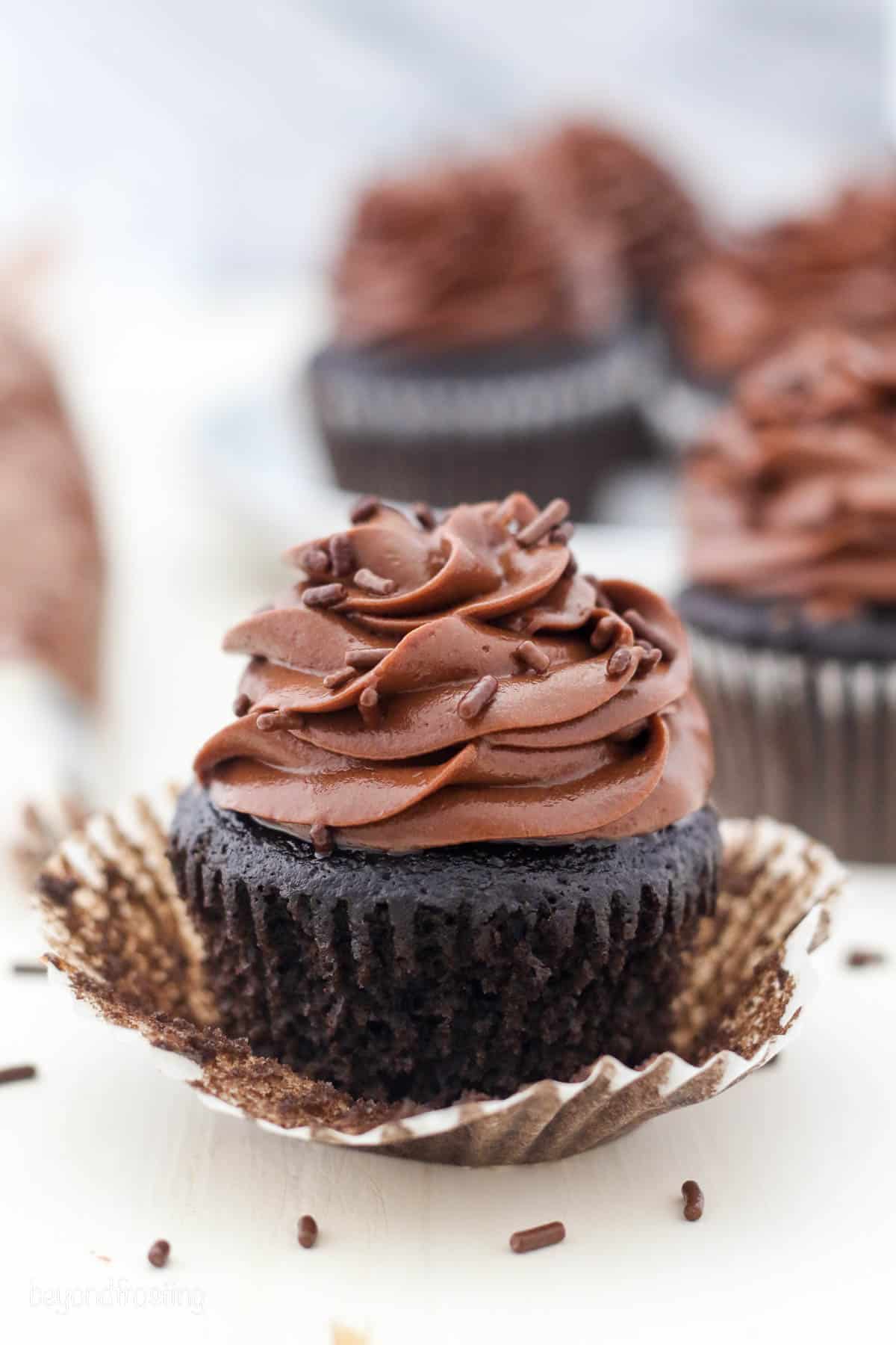 A partially unwrapped chocolate cupcake topped with a swirl of chocolate cream cheese frosting and chocolate sprinkles.