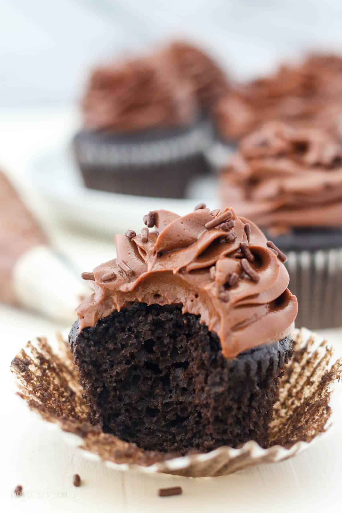 A partially unwrapped chocolate cupcake topped with a swirl of chocolate cream cheese frosting and chocolate sprinkles, with a bite missing.