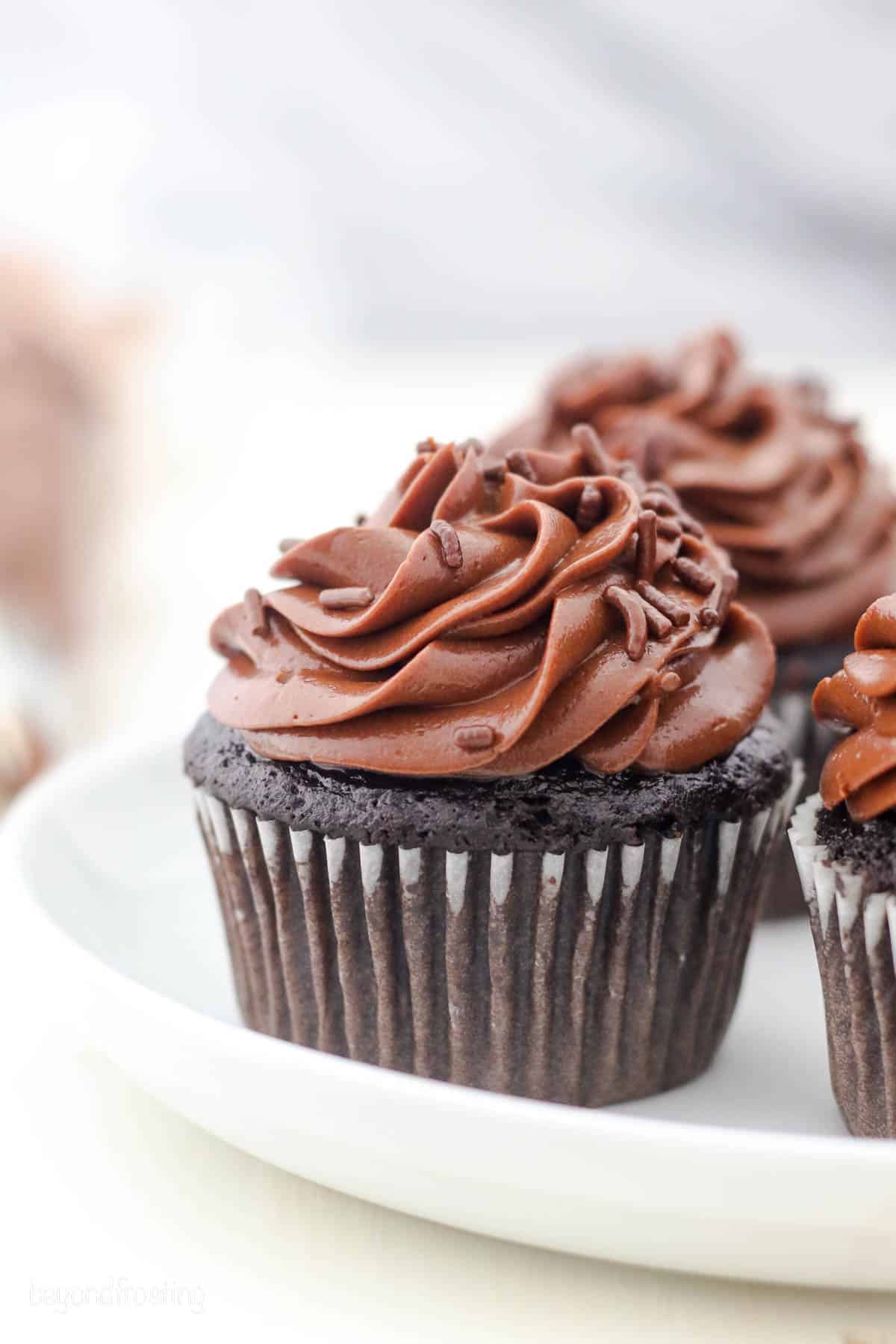 Chocolate cupcakes topped with swirls of chocolate cream cheese frosting and chocolate sprinkles on a white plate.