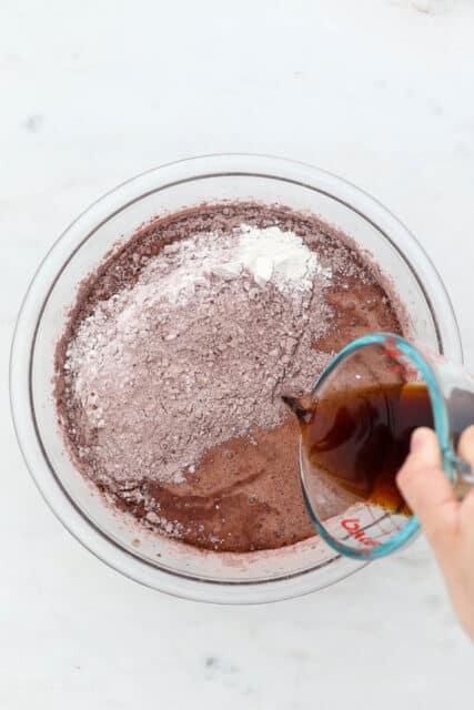 Brewed coffee is poured over cocoa powder and dry ingredients added to chocolate cake batter in a glass bowl.
