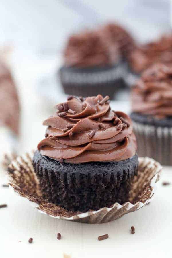 A dark chocolate cupcake is unwrapped sitting on a white board surrounded by sprinkles