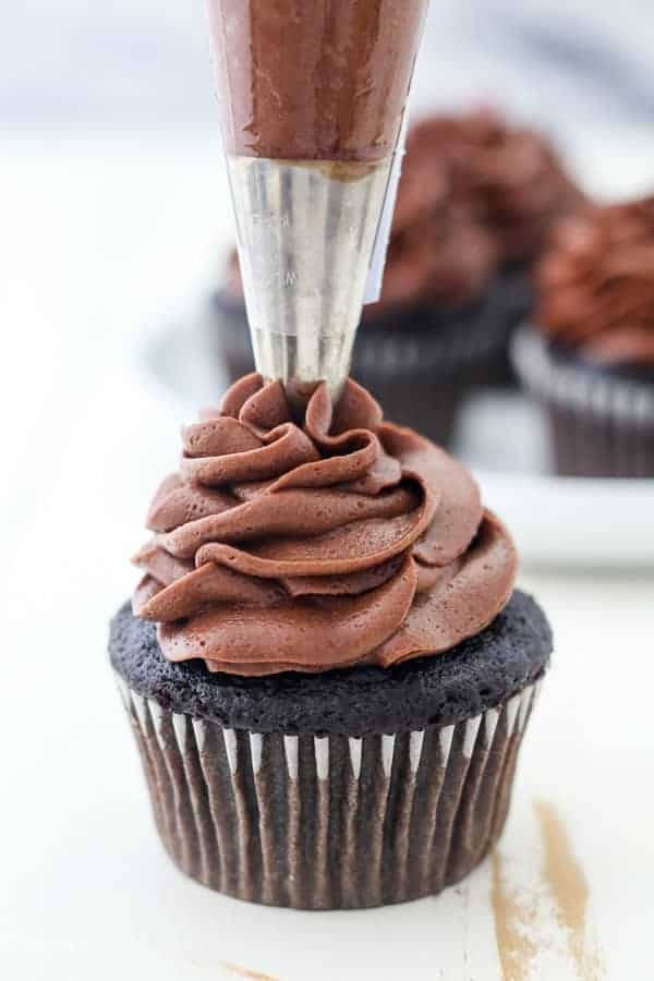 A piping bag fitted with a large piping tip is frosting a dark chocolate cupcake