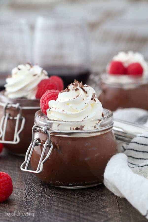 A small jar filled with chocolate pudding topped with whipped cream and raspberries and a glass of red wine blurred out in the background