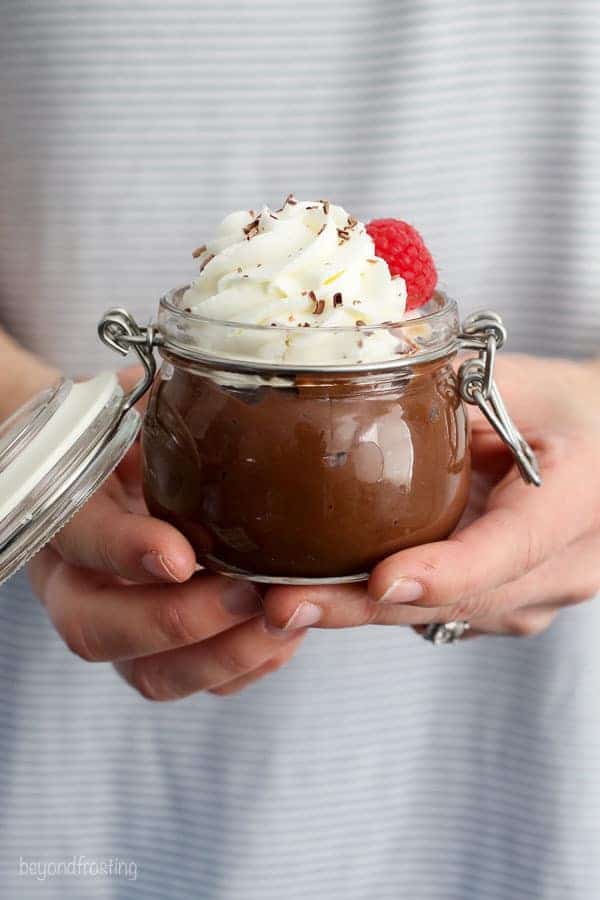 Two hands holding a small glass jar filled with chocolate pudding topped with whipped cream and raspberries