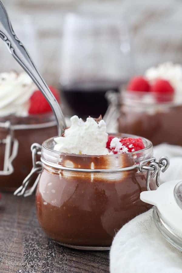 A small jar filled with chocolate pudding topped with whipped cream and raspberries. There's a silver spoon dipped in the jar and it appears to have a couple of bites missing.