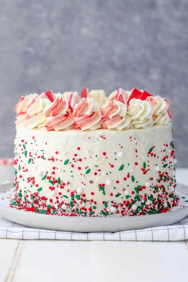 A gorgeous cake frosted with a white buttercream and loads of sprinkles on the outside that are red, green and white. The swirls of buttercream on top are half red and half white and it's garnished with Andes Peppermint Bark Candy.