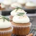 Three gorgeous gingerbread cupcakes with white cupcake wrappers and a rosemary garnish are sitting on a wire baking rack