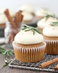 Frosted gingerbread cupcakes on a wire rack topped with small rosemary sprigs.