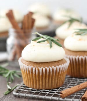 Frosted gingerbread cupcakes on a wire rack topped with small rosemary sprigs.