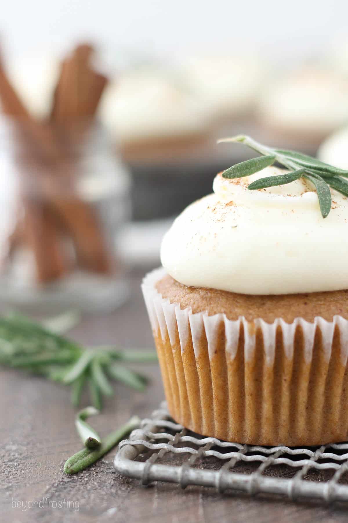 Close up of a frosted gingerbread cupcake on a wire rack topped with a small rosemary sprig.