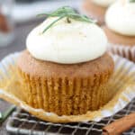 Close up of an unwrapped gingerbread cupcake topped with cinnamon cream cheese frosting and a rosemary sprig.