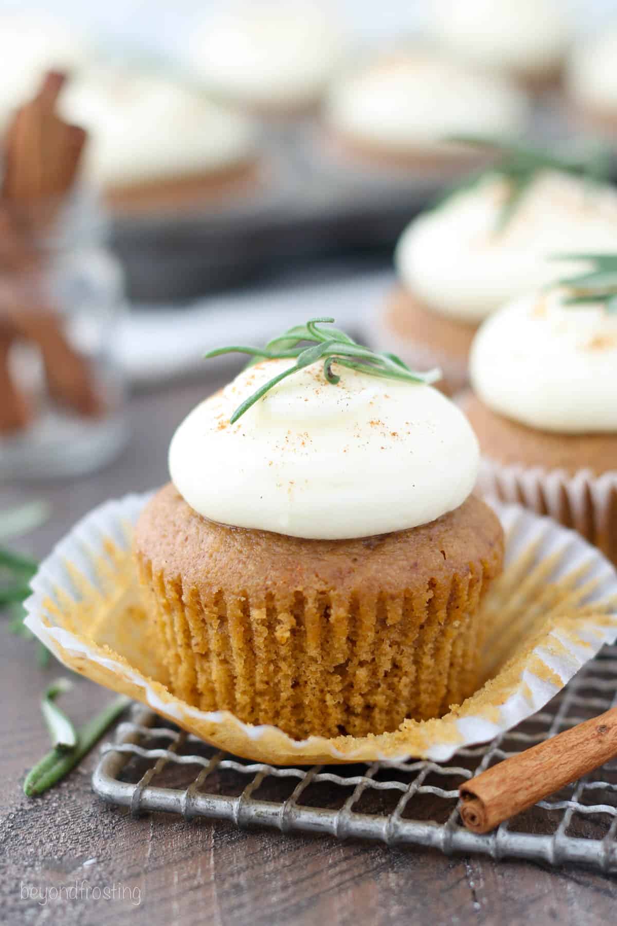 An unwrapped gingerbread cupcake topped with cinnamon cream cheese frosting and a rosemary sprig on a wire rack, with more cupcakes in the background.