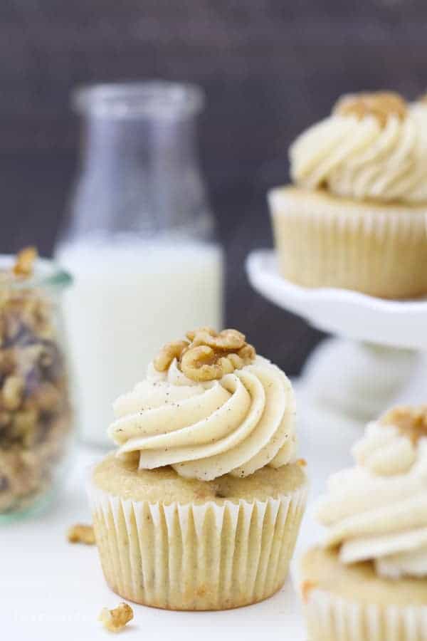 A cupcake is sitting in front of a small glass of milk, a jar of walnuts and a small white cake stand with cupcakes on top.