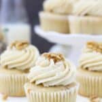 A beautiful vanilla cupcake with a big swirl of frosting on top garnish with a walnut. In the background is a blurred out white cake stand with more cupcakes on top.