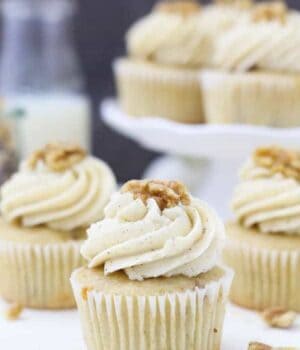 A beautiful vanilla cupcake with a big swirl of frosting on top garnish with a walnut. In the background is a blurred out white cake stand with more cupcakes on top.