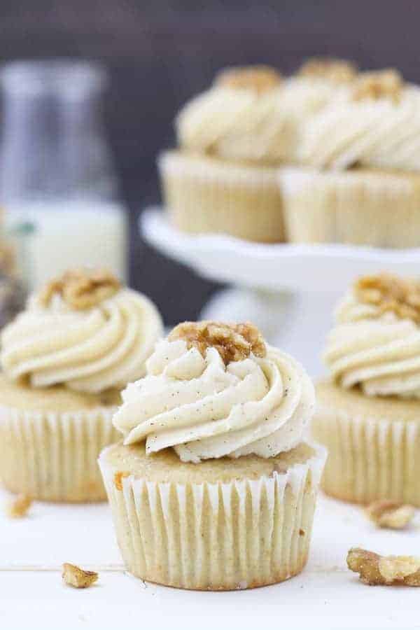 A beautiful vanilla cupcake with a big swirl of frosting on top garnish with a walnut. In the background is a blurred out white cake stand with more cupcakes on top.