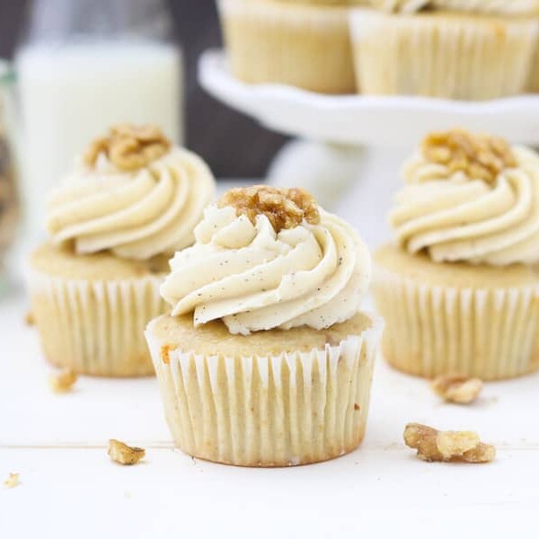 Three maple walnut cupcakes sitting on a white piece of wood with sprinkled walnuts all over.