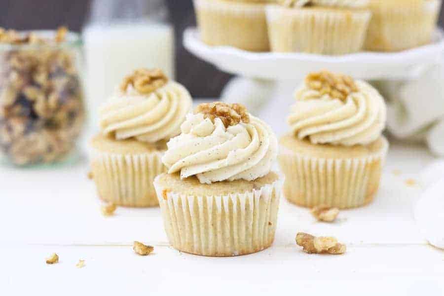 Three maple walnut cupcakes sitting on a white piece of wood with sprinkled walnuts all over.