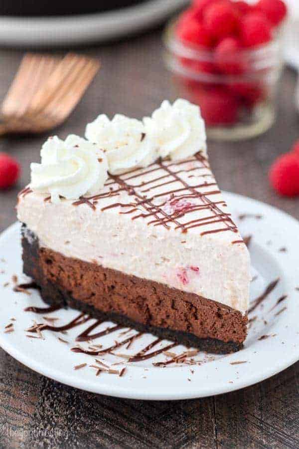 An overhead shot of a slice of raspberry chocolate mousse garnished with a chocolate drizzle and whipped cream with raspberries sprinkled in the background.