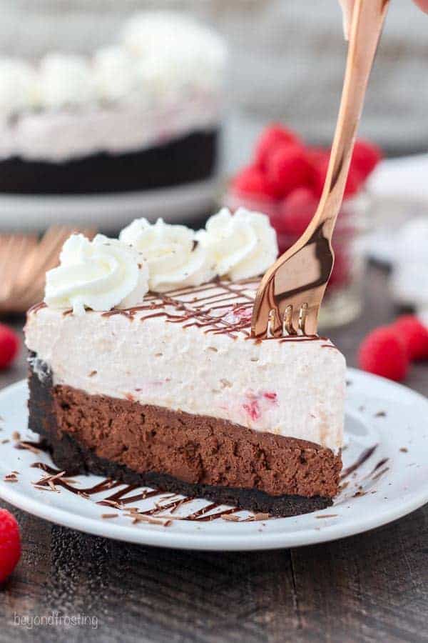 A rose gold fork sinking into a double layer mousse cake with chocolate on the bottom and raspberry on top.