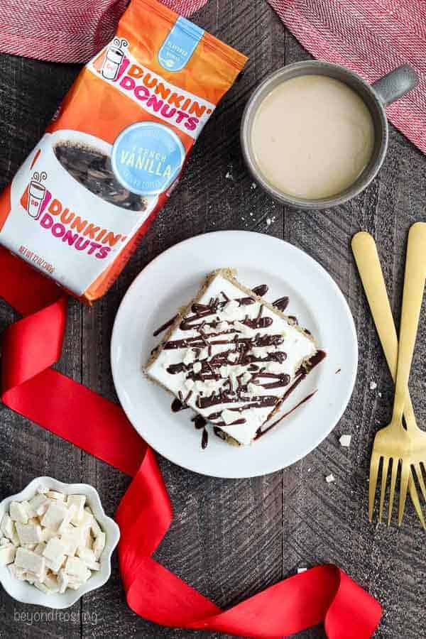 An overhead view of a slice of cake on a white rimmed plate, there's two gold fork, a cup of coffee, a bag of Dunkin Donuts French Vanilla Coffee and a big red ribbon styled on a wooden surface