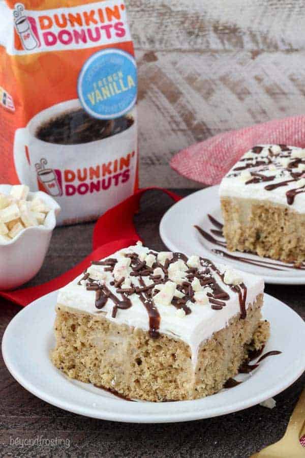 A white plate with a slice of cake covered in whipped cream, fudge and peppermint bark. There's a bag of Dunkin' Donuts French Vanilla coffee in the background.