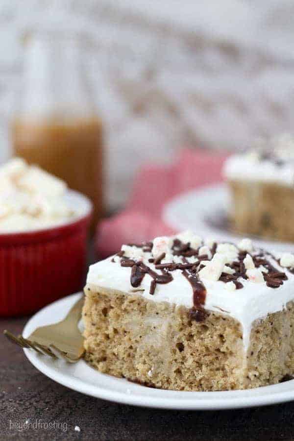 A close up shot of a slice of mocha cake on a white plate with a gold fork resting on the side. The cake is topped with whipped cream, hot fudge sauce and peppermint bark