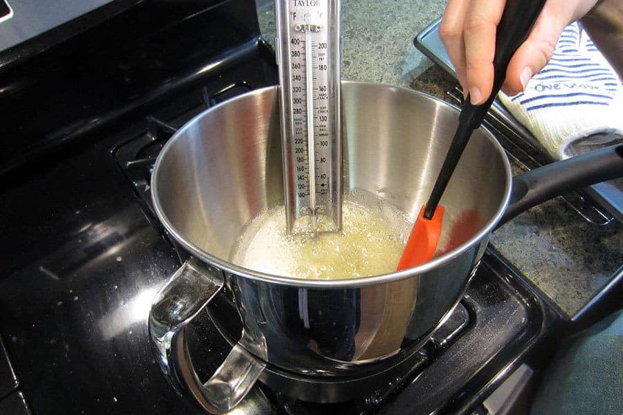 A metal mixing bowl over a pot on the stovetop with a candy thermometer attached