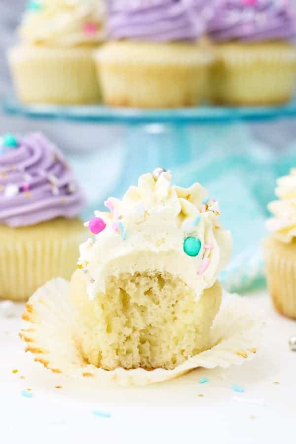 A close up show of a vanilla cupcake topped with Swiss Meringue Buttercream and colorful sprinkles. The cupcake has a big bite taken out of it