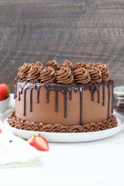 A large chocolate cake on white cake plate. The cake is drizzled with chocolate ganache and covered with frosting rosettes.
