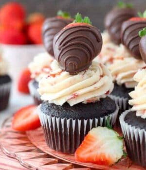 A gorgeous chocolate cupcake with a pretty pink strawberry frosting and a chocolate covered strawberry on top. It's sitting on a pink glass cake plate with a pink striped towel underneath and fresh strawberries scattered around.