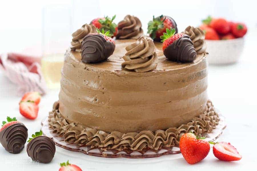 A wide shot of a chocolate layer cake with chocolate frosting, it's garnished with chocolate covered strawberries