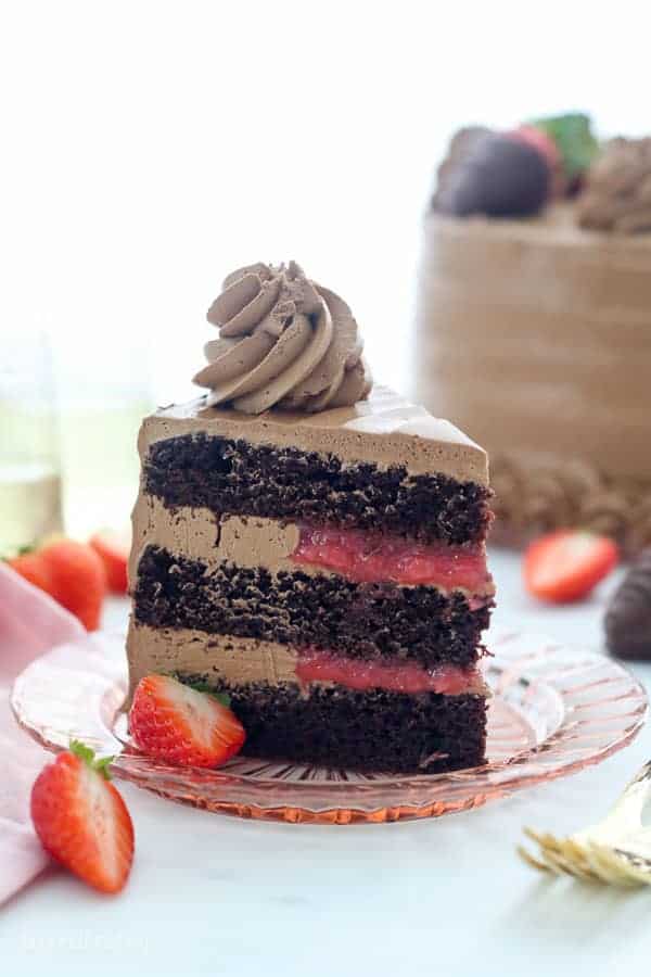 A tall slice of chocolate cake with chocolate frosting and it's filled with a strawberry sauce. The cake is sitting on a pink tinted glass plate and you can see the whole cake blurred out in the background.