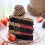 A slice of dark chocolate cake with a strawberry filling and a silky buttercream frosting stands tall on a pink tinted glass plate garnished with strawberries.