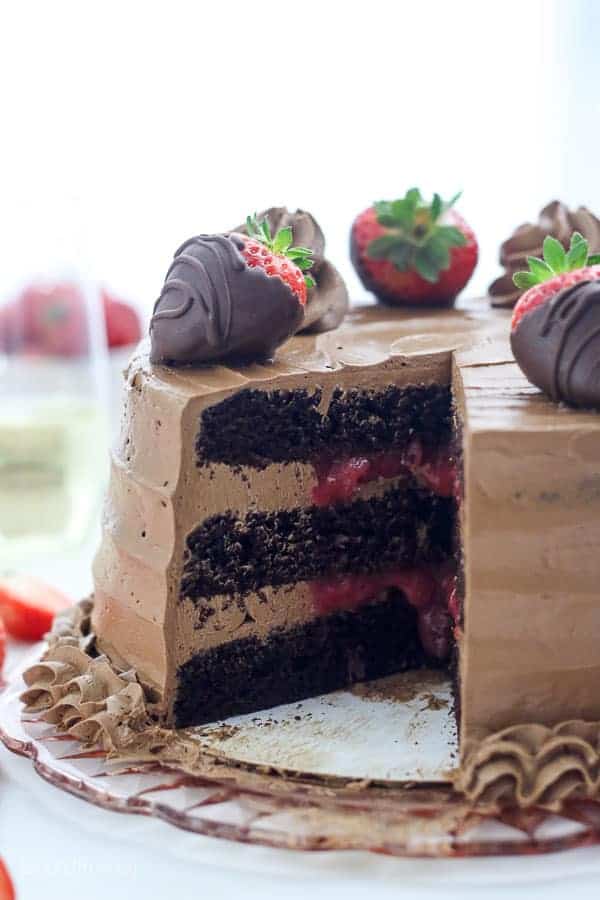 A big chocolate cake with a strawberry filling and a silky buttercream frosting there's a slice missing from the cake revealing the center and it's garnished with chocolate covered strawberries.