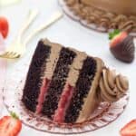 An overhead view of a slice of dark chocolate cake with a strawberry filling and a silky buttercream frosting, there are a few strawberries scattered around and two gold forks blurred out in the background.