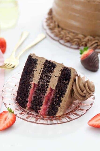 An overhead view of a slice of dark chocolate cake with a strawberry filling and a silky buttercream frosting, there are a few strawberries scattered around and two gold forks blurred out in the background.