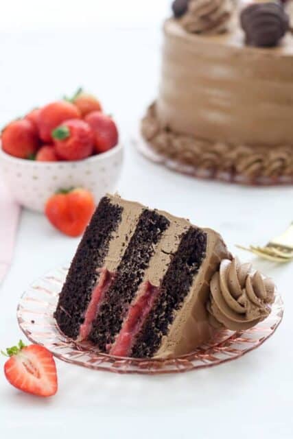 A slice of dark chocolate cake with a strawberry filling and a silky buttercream frosting sitting on a light pink plate, it's surrounded by strawberries