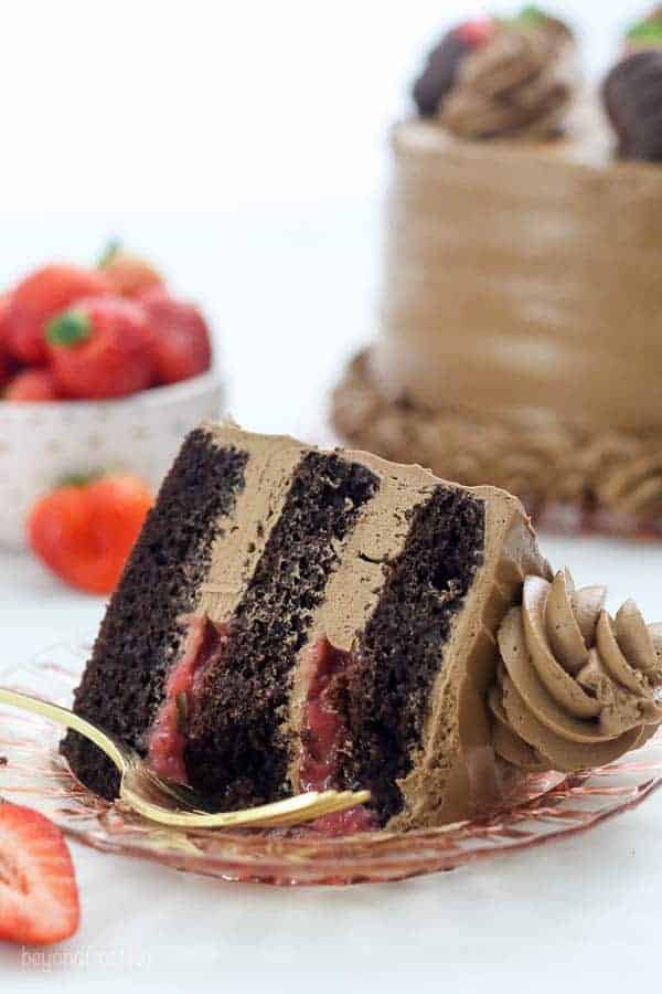 A slice of dark chocolate cake with a strawberry filling and a silky buttercream frosting. The slice of cake is laying on a light pink plate with a gold fork and it's missing a few bites.