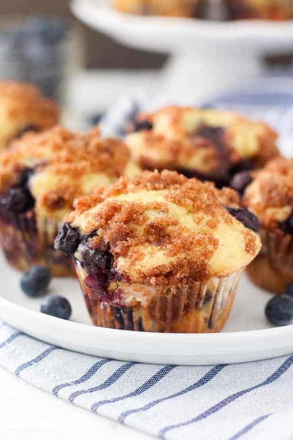 A close up shot of a blueberry muffins on a white rimmed plate which is sitting on a blue and white stripped napkin. There's a cinnamon sugar crumb topping.