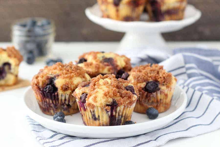 A round white rimmed plate with 4 blueberry muffins with a crumb topping.
