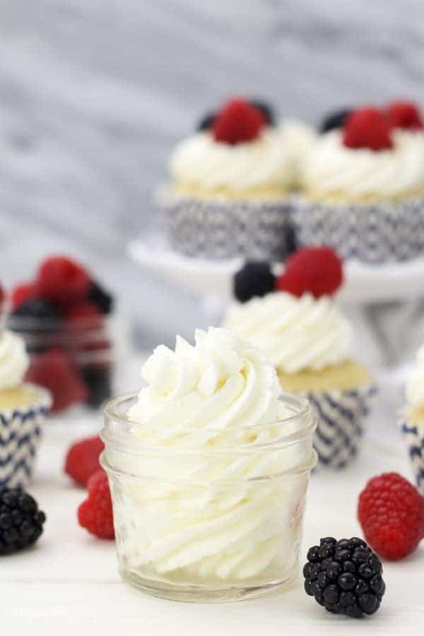 A mini mason jar filled with a gorgeous swirl of mascarpone whipped cream, there are cupcakes in the background with the same frosting and covered with berries.