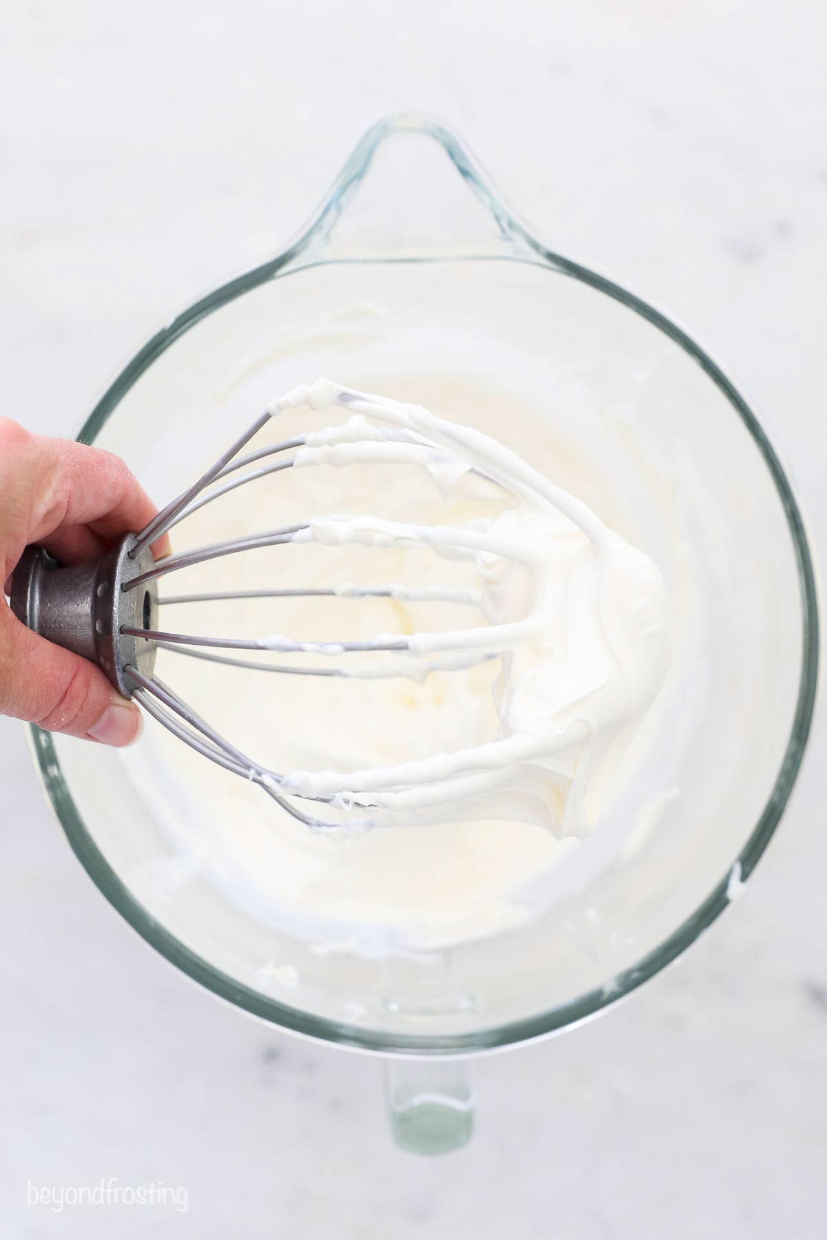 A hand holding a stand mixer attachment covered with mascarpone whipped cream over a mixing bowl.