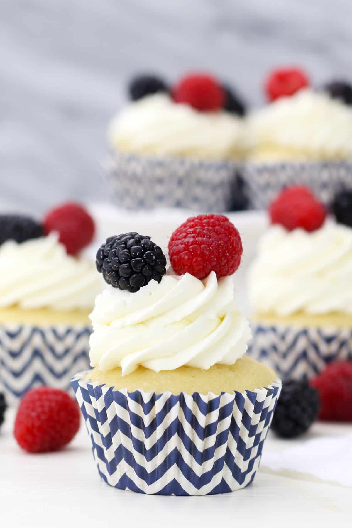 A cupcake in a blue and white striped cupcake liner frosted with a swirl of mascarpone whipped cream and topped with fresh berries, with more frosted cupcakes in the background.