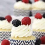 A close up of a vanilla cupcake in a navy blue chevon striped cupcake liner with a whipped frosting and a blackberry and raspberry on top