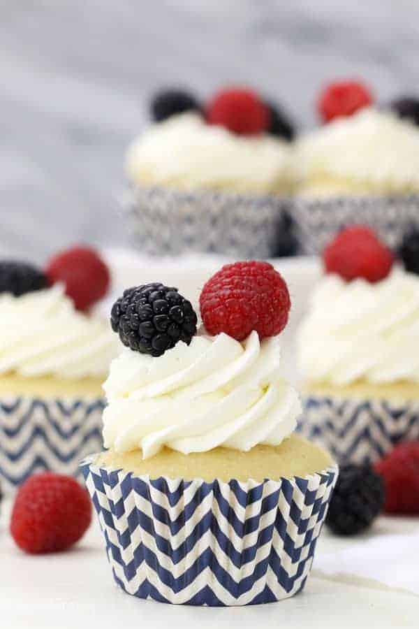 A close up of a vanilla cupcake in a navy blue chevon striped cupcake liner with a whipped frosting and a blackberry and raspberry on top