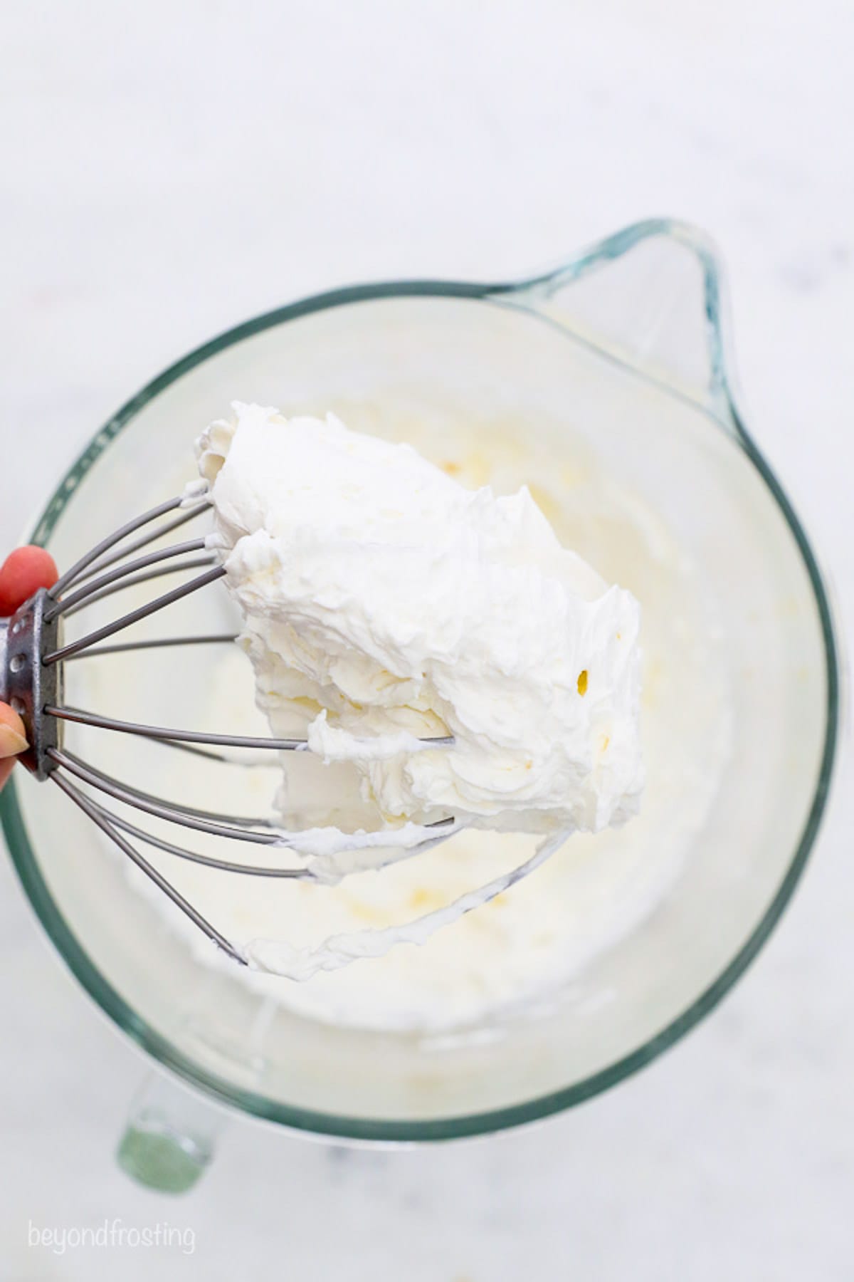 A hand holding a stand mixer attachment covered with mascarpone whipped cream frosting over a mixing bowl.