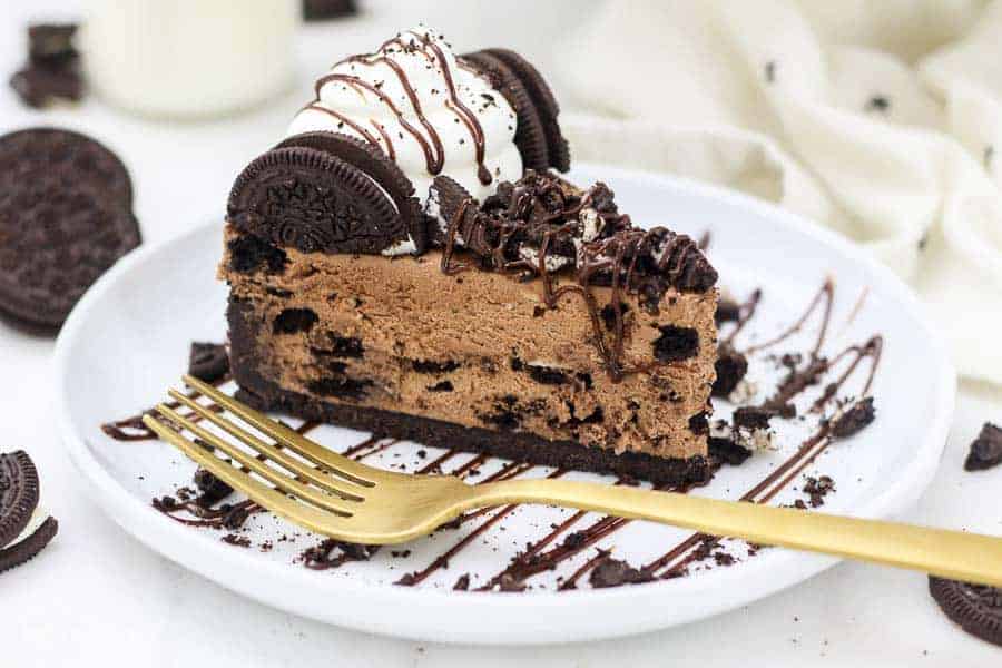 a horizontal image shows a big slice of chocolate Oreo cheesecake on a white rimmed plate with a gold fork hanging off the side. The cheesecake is loaded with Oreos and topped with whipped cream and chocolate sauce.